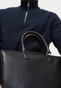 Czarna skórzana torebka z podwójnymi okrągłymi uchwytami i srebrnymi detalami, przedstawiona na tle fakturowanego, ciemnego swetra.