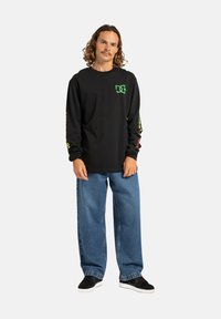 Black long-sleeve t-shirt with green logo, bright graphics on sleeves. Paired with loose blue denim jeans and black sneakers.