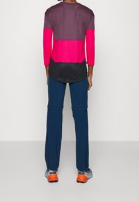 Flerfärgad träningsjacka med lång ärm, med lila, rosa och svarta sektioner. Bärs med marinblå byxor och skor med orange detaljer.