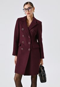 Cappotto bordeaux doppiopetto con grandi bottoni in tonalità argento, tasche laterali e una vestibilità su misura. Il modello tiene in mano una borsa nera.