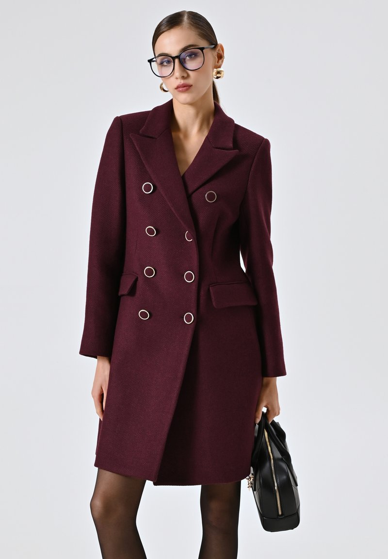 Cappotto bordeaux doppiopetto con grandi bottoni in tonalità argento, tasche laterali e una vestibilità su misura. Il modello tiene in mano una borsa nera.