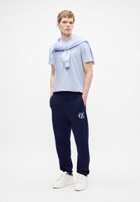 Világoskék pamut póló, laza tengerészkék joggingnadrág logóval, és fehér sportcipő. A póló a vállaknál megkötve, azonos anyaggal.