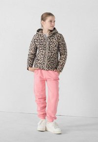 Gewatteerde jas met luipaardprint, capuchon, ritssluiting en beige achtergrond. Gecombineerd met roze joggingbroek en witte sneakers.