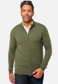 Cardigan zippé en vert olive, en matériau tricoté doux. Présente un col montant et des poignets côtelés. porté sur une chemise blanche.