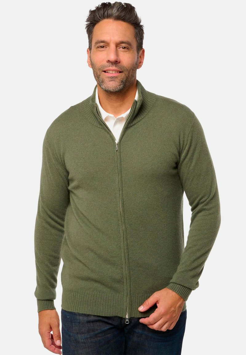 Cardigan zippé en vert olive, en matériau tricoté doux. Présente un col montant et des poignets côtelés. porté sur une chemise blanche.