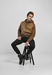Brun hettegenser med teksturert front og ensfarget bak, parret med svarte tettsittende bukser og svarte snørelester, sittende på en krakk.