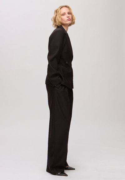 Femme blonde debout, les mains dans les poches, portant un costume noir avec un pantalon à jambes larges et des chaussures noires pointues sur un fond uni.