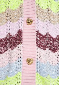 Neulejakku, jossa on aaltoileva kuvio vihreän, vaaleanpunaisen, ruskean ja sinisen sävyissä, ja ribbaus vaaleanpunaisella kauluksella sekä kultaisilla sydämenmuotoisilla napeilla.