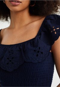 Marineblauwe top met een gegolfde oogjesrand bij de hals, voorzien van bloemen uitgesneden. De stof is gathered met een textuur smock-design.