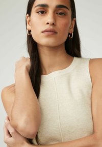 Beige ribbet tanktop med rund halsudskæring. Har en subtil tekstur. Guld hoops og naturligt hår supplerer looket.