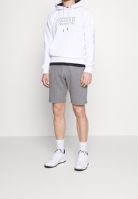 Sweat à capuche blanc avec un logo gris, associé à un short gris et des baskets blanches. La tenue est décontractée et comporte une capuche avec cordon de serrage.