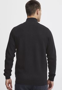 Svart stickad tröja med hög krage, långa ärmar och ribbade cuffar. Slät textur med en figurnära design, inga synliga mönster eller utsmyckningar.
