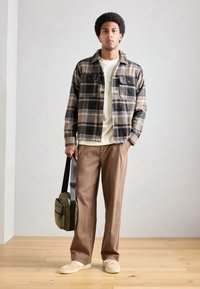 Plaid flannel shirt in beige and black, beige t-shirt, brown trousers, and tan sneakers. Green bag with black strap, wooden floor background.
