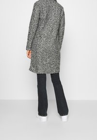 Grå och svart spräcklig lång kappa med rak skärning, textil med struktur och klassisk krage, kombinerad med mörk, utsvängd jeans och vita skor.