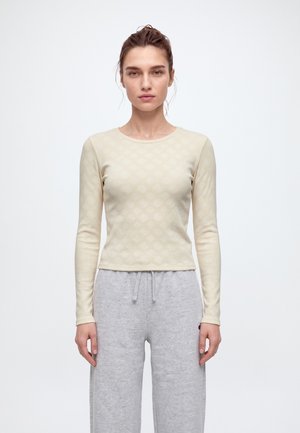 Une femme se tient face à l’objectif, vêtue d’un haut crème à manches longues et d’un pantalon de jogging gris clair à cordon, sur un fond uni.