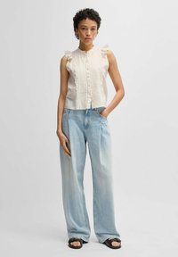 Ljusbeige ruffle ärmlös topp med fackdetaljer och knappar, kombinerad med vida blekta blå jeans och svarta flip-flops.