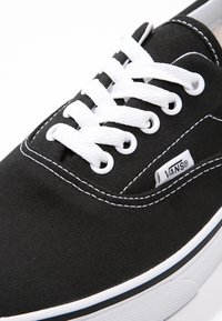 Gros plan d'une basket Vans noire avec des lacets blancs, des coutures blanches et une semelle en caoutchouc blanche avec des rayures bleues et noires.