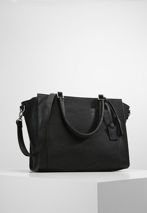 Sac fourre-tout en cuir noir avec double poignées, une bandoulière amovible et un petit pompon en accent. Texture lisse, forme structurée.