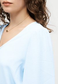 Γυναίκα με καστανά σγουρά μαλλιά που φοράει ένα ανοιχτό μπλε υφασμάτινο τοπ με V-λαιμόκοψη και ένα λεπτό χρυσό κολιέ με μικρό κρεμαστό.