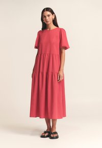 Une robe mi-longue à plusieurs volants de couleur corail vif, confectionnée dans un tissu léger, avec des manches courtes bouffantes et un col rond. Sandales noires.