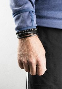 Black leather and bead bracelets on a wrist, layered with textured patterns, against a blue sweatshirt and black pants background.