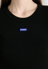 Gros plan d'une personne portant un t-shirt noir côtelé avec un petit patch bleu intitulé "HUGO" sur la zone de la poitrine.