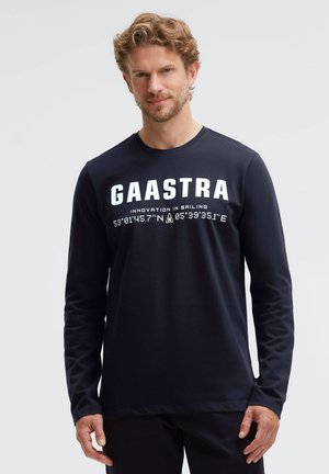 Man with light brown hair and beard wearing a black long-sleeve shirt with "GAASTRA Innovation in Sailing" and coordinates printed in white.