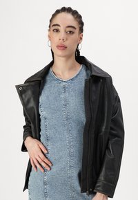 Casaco de couro preto com um design clássico, apresentando um colarinho largo e fecho de correr completo, sobre um vestido de ganga azul claro.