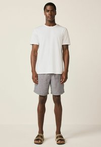 Junger Mann steht, trägt ein schlichtes weißes T-Shirt, graue Shorts und beige Sandalen vor einem neutralen Hintergrund.