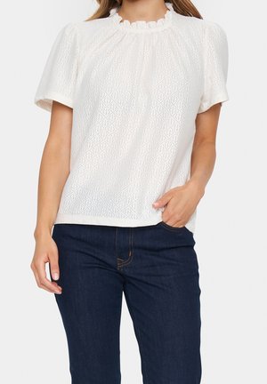 Kobieta ubrana w białą, krótkim rękawem, ażurową bluzkę z falbanką przy dekolcie oraz ciemnoniebieskie dżinsy, z ręką w kieszeni, na jednolitym tle.