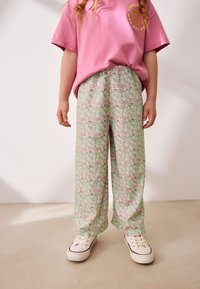 T-shirt rose à manches courtes avec un motif de soleil, associée à un pantalon ample vert à motifs floraux roses et violets, portée avec des baskets blanches.