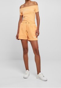 Persikka olkapäältä avattava toppi lyhyillä hihoilla, yhdistettynä yhteensopivaan joustavaan vyötärölahkeisiin shortseihin, joissa on sivutaskut ja hillitty logo. Valkoiset lenkkarit.