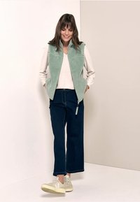 Ljusgrön fleeceväst med dragkedja i fronten, tillsammans med en krämfärgad topp och mörk denimvidbyxor. Skorna är ljusbeige sneakers.