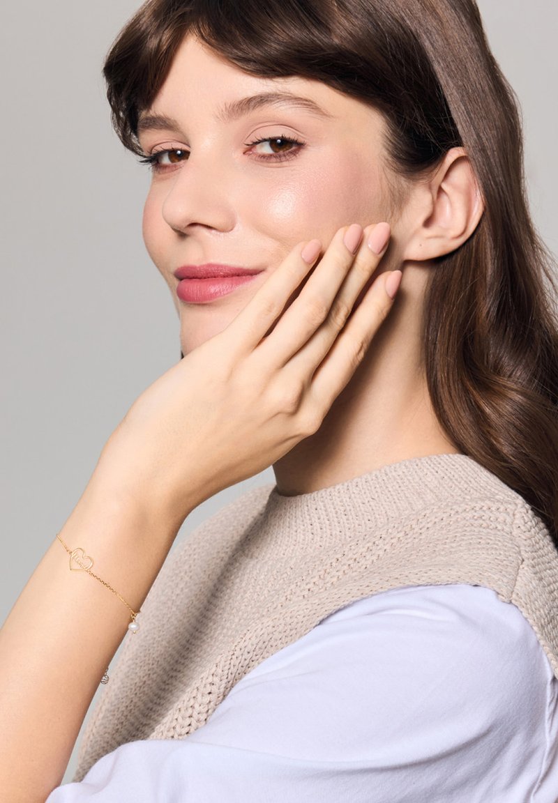 Złota bransoletka z zawieszką w kształcie serca i perłowym akcentem. Miękki, jasnoszary sweter dzianinowy z okrągłym dekoltem i białym rękawem pod spodem.