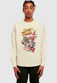 Cream-colored sweatshirt featuring a multicolored "Street Fighter II" graphic with characters in action poses. Long sleeves and relaxed fit.