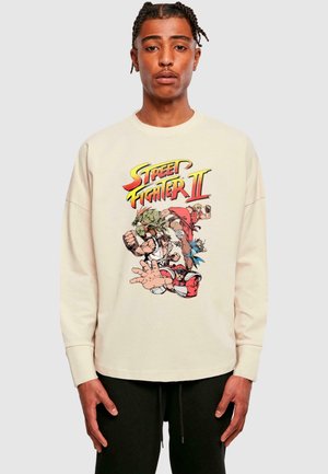 Cream-colored sweatshirt featuring a multicolored "Street Fighter II" graphic with characters in action poses. Long sleeves and relaxed fit.