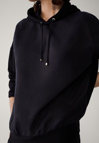 Zwarte hoodie gemaakt van gladde stof met een losse pasvorm, voorzien van een trekkoord en metalen accenten bij de halslijn.