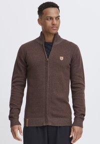 Brauner Reißverschluss-Strickcardigan mit strukturiertem Strick, gerippten Bündchen und Kragen. Mit einem kleinen Emblem auf der Brust und einem Stoffetikett am Saum.