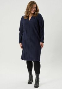 Tumedast sinise värviga kudumkleit V-kaelusega, tekstuurse leevenduskudumisega, pikkade käistega ja põlvepikkuses, koos mustade retuuside ja turske pahkluu saapaga.