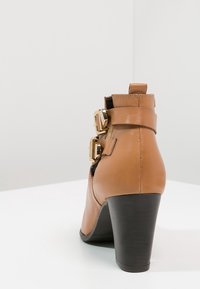 Bottines en cuir tan avec deux boucles dorées et un haut talon empilé noir, montrées de l'arrière sur une surface et un fond blancs.