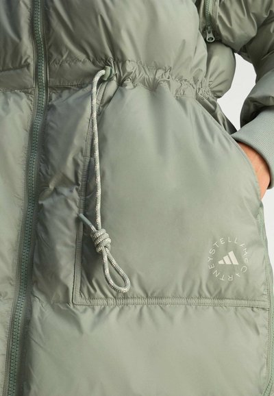 Primer plano de una chaqueta acolchada verde claro que cuenta con un cordón ajustable en la cintura y un logo circular en el bolsillo.
