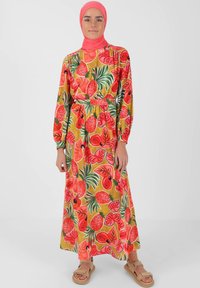 Maxime dress in bright yellow-green fabric with a colorful fruit and leaf pattern. Long sleeves, belted waist, and a loose fit. Pink headscarf.