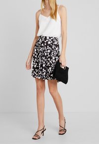 Černá květinová sukně s bílým vzorem, spojená s bílým tílkem bez rukávů a černou kabelkou. Na nohou má páskové černé podpatky.