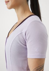 Haut à manches courtes violet clair avec une texture côtelée, un décolleté en V et des accents violet foncé le long du col. Logo affiché sur la manche.