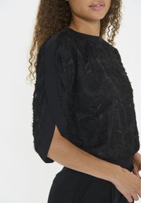 Black top with textured, sheer fabric featuring a floral pattern. Short sleeves and a round neckline. Lightweight and smooth material.