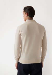 Polo shirt bej cu mânecă lungă, cu guler și tiv ribbotat, având o textură netedă și un design ajustat.