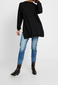 Felpa nera oversized con maniche lunghe, caratterizzata da un collo rotondo e polsini a costine, abbinata a jeans chiari strappati e stivali neri alla caviglia.