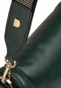 Primo piano di un angolo di una borsa in pelle verde scuro con chiusura dorata e tracolla staccabile in tessuto intrecciato bianco e nero con etichetta dorata.