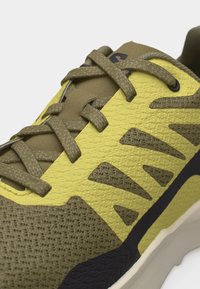 Κοντινό πλάνο αθλητικού παπουτσιού σε ελιά πράσινο και κίτρινο χρώμα με υφή υφάσματος, διακοσμητικά σχέδια και κορδόνια σε ταιριαστό χρώμα σε λευκό φόντο.