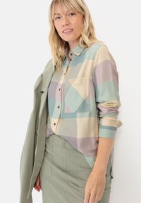 Button-up shirt featuring a large check pattern in pastel colors of blue, beige, and purple, with a single chest pocket and rolled sleeves.